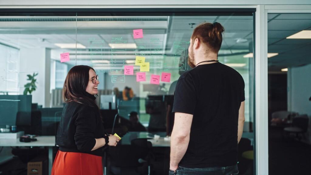 Codemate’s designers Tiina and Olli brainstorming and working on the project task list at the office. Tiina smiles during the conversation, and colourful post-it notes are attached to the wall, serving as brainstorming tools.