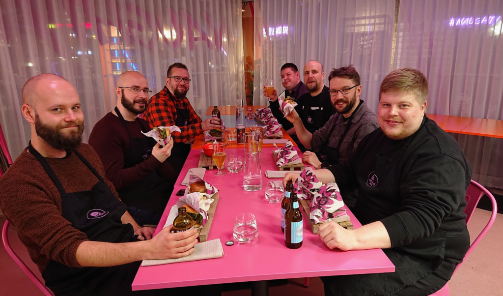 Seven man around the pink table eating hamburgers and having drinks.