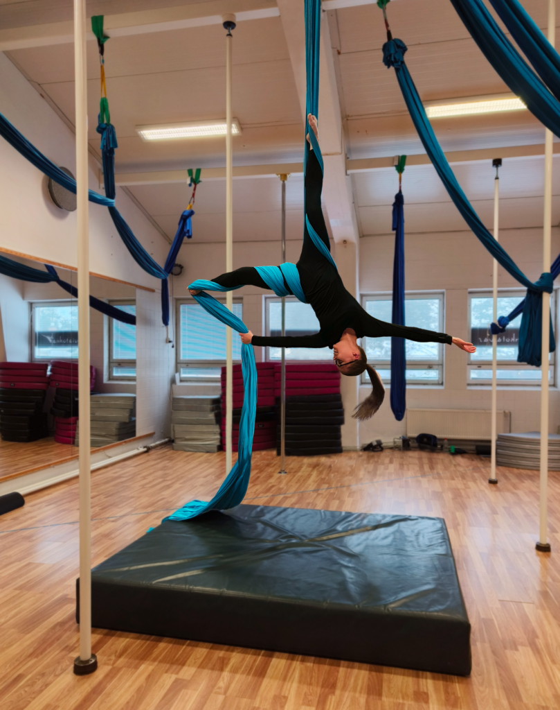 A woman is performing an aerial silk routine in a gymnasium, suspended upside down with blue silks wrapped around her legs and waist. She is wearing a black bodysuit and extends one arm gracefully. The background features mirrors, gym mats, and multiple aerial silks hanging from the ceiling.