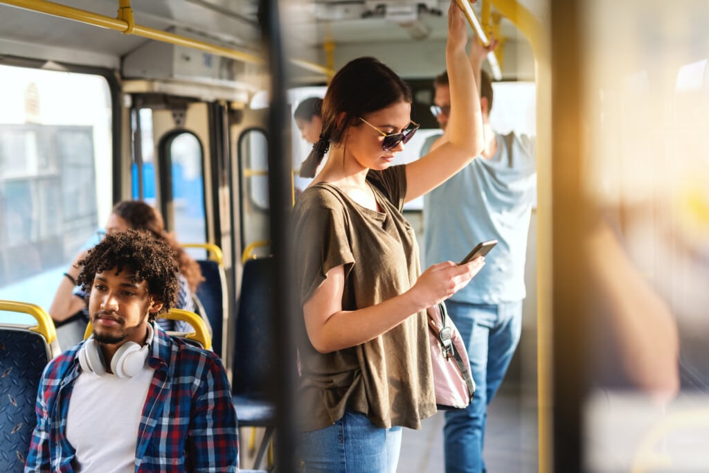 Nainen seisoo bussissa pidellen kaiteesta ja katsoo puhelintaan aurinkolasit päässään. Hänellä on rento t-paita ja olkalaukku. Taustalla näkyy muita matkustajia, kuten istuva mies, jolla on kuulokkeet kaulassa, sekä bussin sisätiloja ja ikkunoista heijastuvaa valoa.