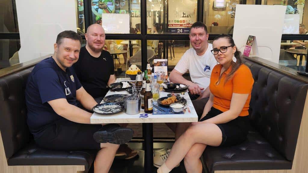 Four people are sitting in a restaurant booth, smiling at the camera. The table is filled with plates, bottles of beer, and desserts, including mango sticky rice. The setting is a casual dining restaurant with a glass window in the background, showing another seating area. The group appears to be enjoying a meal together.